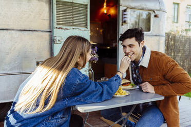 Seitenansicht eines jungen Mannes und einer Frau, die sich lächelnd anschauen, während sie ein köstliches romantisches Abendessen in der Nähe eines schäbigen Wohnwagens genießen - ADSF03714