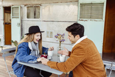 Junger Mann und Frau lächeln und schauen sich an, während sie an einem Tisch mit einem heißen Getränk und einem Stück Geburtstagskuchen in der Nähe eines verwitterten Wohnmobils sitzen - ADSF03720