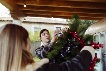 Young man and woman in stylish clothes decorating lovely Christmas tree near aged caravan in countryside - ADSF03729