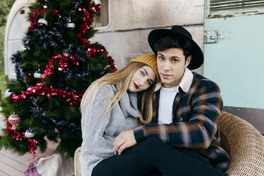 Fröhlicher junger Mann und Frau, die sich gegenseitig umarmen, während sie in einem Sessel in der Nähe eines schönen Weihnachtsbaums vor einem alten Lieferwagen sitzen - ADSF03744