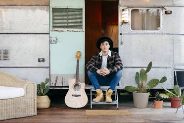 Gut aussehender junger Mann in stilvollem Outfit, der lächelnd in der Tür eines alten Wohnwagens neben einer Akustikgitarre sitzt - ADSF03745