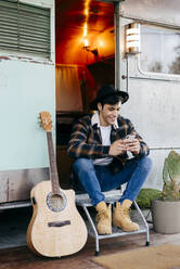Gut aussehender junger Mann in stilvollem Outfit, der lächelt und ein modernes Smartphone benutzt, während er in der Tür eines alten Wohnwagens neben einer akustischen Gitarre sitzt - ADSF03746