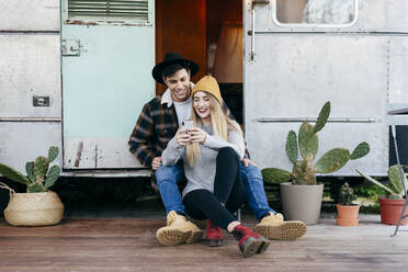 Attractive young man and woman smiling and browsing modern smartphone while sitting near shabby camper together - ADSF03748