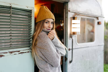 Charming young female in trendy outfit smiling and looking at camera while standing in entrance of shabby camper - ADSF03754