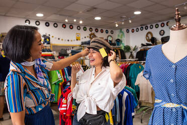 Zwei reizende Frauen lachen und probieren einen schönen Hut an, während sie Zeit in einem kleinen Laden miteinander verbringen - ADSF03772