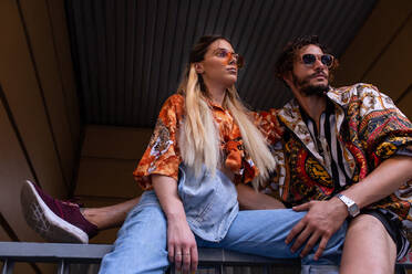 Junger Mann und Frau in trendigen Outfits sitzen auf einem Metallzaun auf einer Straße und schauen in die Kamera - ADSF03790