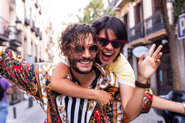 Handsome bearded man in trendy outfit smiling and giving piggyback ride to cheerful girlfriend on blurred background of city street - ADSF03793