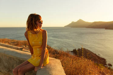 Beautiful woman in stylish short dress and sunglasses sitting on terrace with amazing view of seascape in sunset - ADSF03806