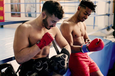 Ethnische und weiße muskulöse Männer sitzen in einem Boxclub und ziehen sich Handschuhe an. - ADSF03852