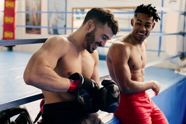 Ethnische und weiße muskulöse Männer sitzen in einem Boxclub und ziehen sich Handschuhe an. - ADSF03853
