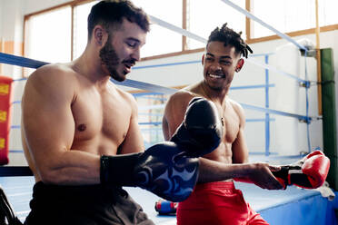 Ethnische und weiße muskulöse Männer sitzen in einem Boxclub und ziehen sich Handschuhe an. - ADSF03854