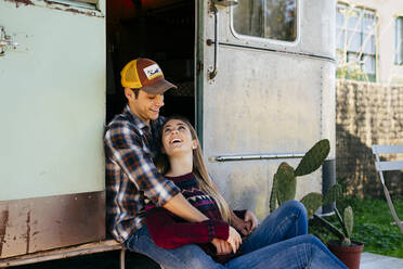Gut aussehender junger Mann umarmt hübsche Frau von hinten, während sie zusammen in der Nähe eines schäbigen Wohnwagens an einem sonnigen Tag sitzen - ADSF03867