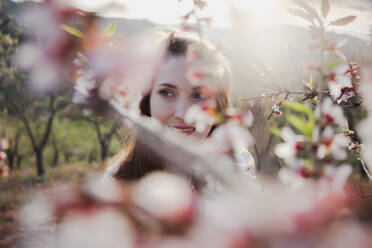 Blick durch die Zweige des blühenden Waldes auf eine attraktive, fröhliche Dame, die im Garten zwischen Hügeln wegschaut - ADSF03932