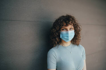 Brunette curly woman wearing protective mask in front of grey wall - OYF00157