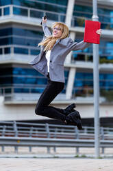 Beautiful blond with glasses in gray jacket and black jeans smiling and leaping with red bag and phone in hands - ADSF04057