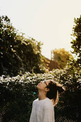 Seitenansicht einer hübschen jungen Frau mit Pullover und Brille, die nach oben schaut, während sie in der Nähe von Büschen im Garten steht - ADSF04084
