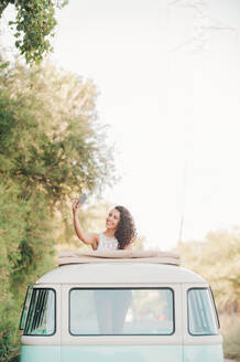 Lovely young female smiling and posing for selfie while standing in hatch of vintage van during trip in nature - ADSF04110