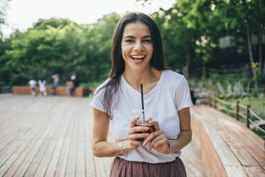 Fröhliche junge Frau, die einen Becher mit Softdrinks hält, während sie im Park steht - OYF00161