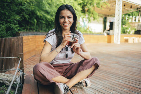 Lächelnde junge Frau, die ein Erfrischungsgetränk in der Hand hält und auf einer Bank im Park sitzt - OYF00164