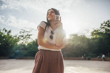 Smiling woman holding soft drink looking away while standing against sky in park - OYF00166