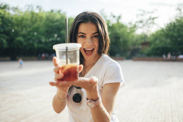 Cheerful young woman with mouth open holding soft drink cup while standing in park - OYF00168