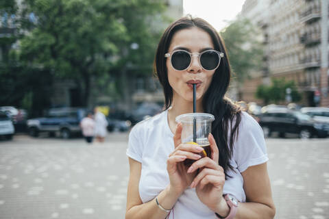 Junge Frau mit Sonnenbrille, die ein Erfrischungsgetränk trinkt, während sie in der Stadt steht, lizenzfreies Stockfoto