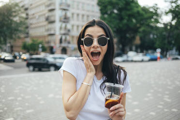 Cheerful woman wearing sunglasses with mouth open standing in city - OYF00171