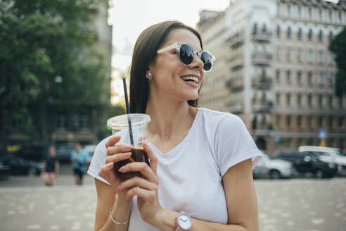 Fröhliche schöne Frau mit Sonnenbrille, die ein Erfrischungsgetränk hält, während sie in der Stadt steht - OYF00172