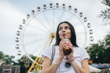 Junge Frau, die ein Erfrischungsgetränk trinkt, während sie an einem Riesenrad in einem Vergnügungspark steht - OYF00174