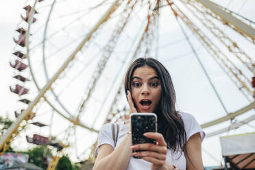 Überrumpelte junge Frau mit offenem Mund, die ihr Smartphone gegen ein Riesenrad im Vergnügungspark hält - OYF00178