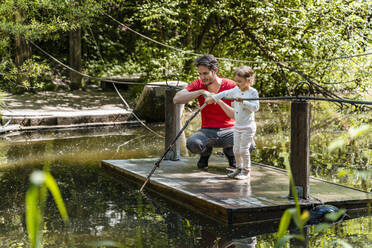Mädchen spielt mit Stock, während sie mit ihrem Vater auf einem Holzfloß über einem See im Wald steht - DIGF12762