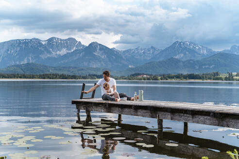 Father with daughter sitting on jetty over lake against mountains - DIGF12764