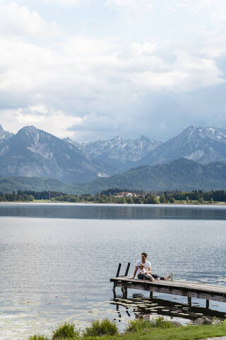 Vater mit Tochter sitzt auf dem Steg über dem See gegen Berge und bewölkten Himmel, lizenzfreies Stockfoto