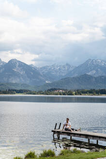 Father with daughter sitting on jetty over lake against mountains and cloudy sky - DIGF12765