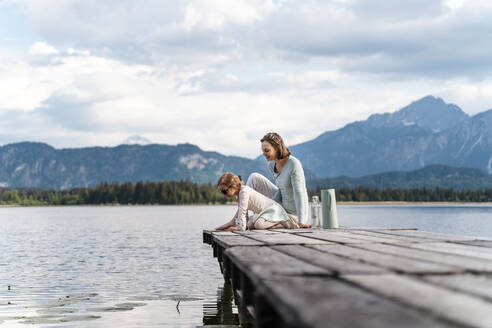 Mother with daughter sitting on jetty over lake against sky - DIGF12766