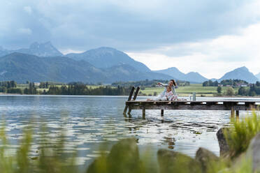 Mutter mit Tochter, die auf einem Steg über dem See sitzen und die Aussicht gegen den Himmel betrachten - DIGF12767