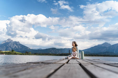 Mother and daughter sitting on jetty over lake against cloudy sky - DIGF12768