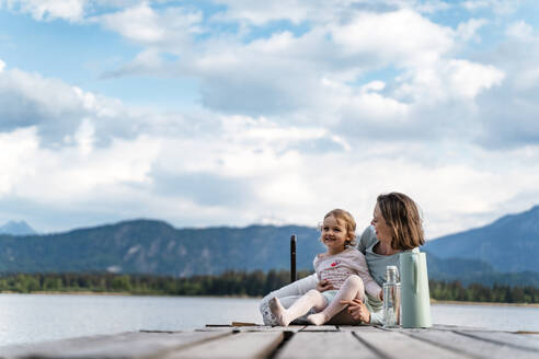 Happy mother and daughter sitting with bottles on jetty against cloudy sky - DIGF12769