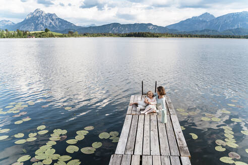 Mid adult woman and daughter sitting on jetty over lake against sky - DIGF12770