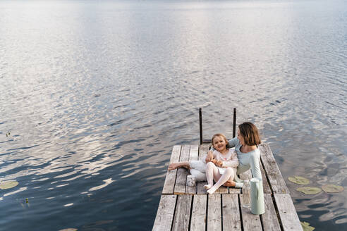 Mid adult woman and daughter relaxing on jetty over lake - DIGF12771