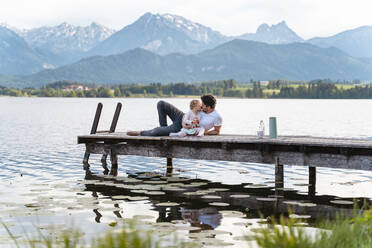Father kissing daughter while sitting on jetty over lake against mountains - DIGF12773
