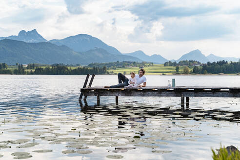 Father and daughter looking at view while relaxing on jetty over lake against mountains - DIGF12774