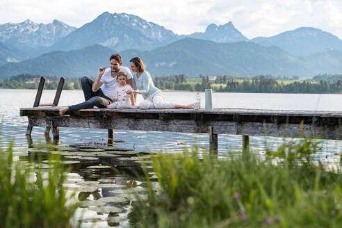 Parents with daughter sitting on jetty over lake against mountains - DIGF12775