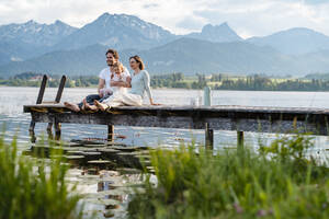 Happy family looking at view while sitting on jetty over lake against mountains - DIGF12776