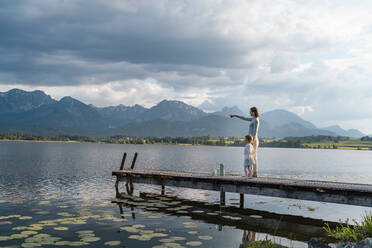 Mother pointing while standing with daughter on jetty over lake against cloudy sky - DIGF12778