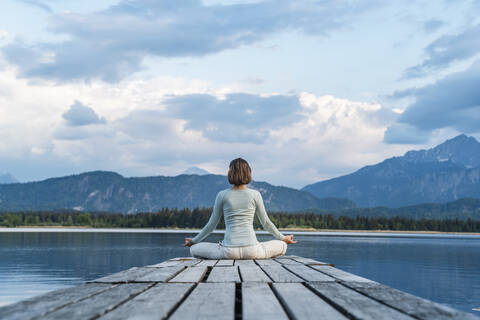 Mid adult woman meditating while sitting on jetty over lake against cloudy sky stock photo