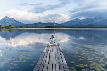 Frau meditiert auf einem Steg über einem See vor bewölktem Himmel sitzend - DIGF12786