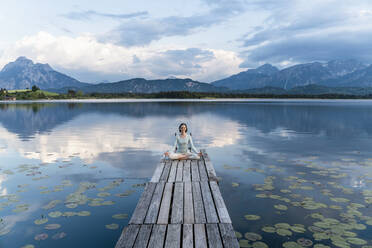 Woman meditating while sitting on jetty over lake against cloudy sky - DIGF12786