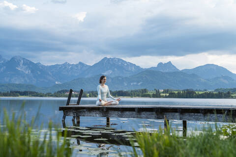 Mittlere erwachsene Frau meditiert auf einem Steg über einem See vor bewölktem Himmel, lizenzfreies Stockfoto