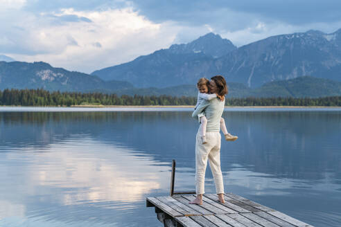 Glückliche Mutter, die ihre Tochter trägt, während sie auf einem Steg über einem See vor einem Berg steht - DIGF12790